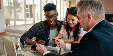 Sitting at a table in a conference room, a realtor in a dark suit sits next to a young man and woman, the woman in a a white shirt and sunglasses with her arms crossed as she smiles. The man in a blue and red flannel shirt and glasses, smiling as he looks at the document the realtor is holding. A computer sits off to the left side of them.