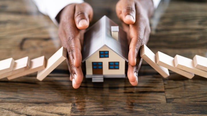 Close up of a Black person's hands on the sides of a miniature house, keeping barriers at bay.