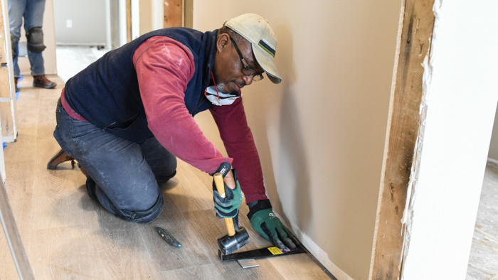 A man kneeling on the ground to install flooring in a home.