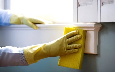 A person wearing yellow cleaning gloves scrubbing a window sill with a sponge.